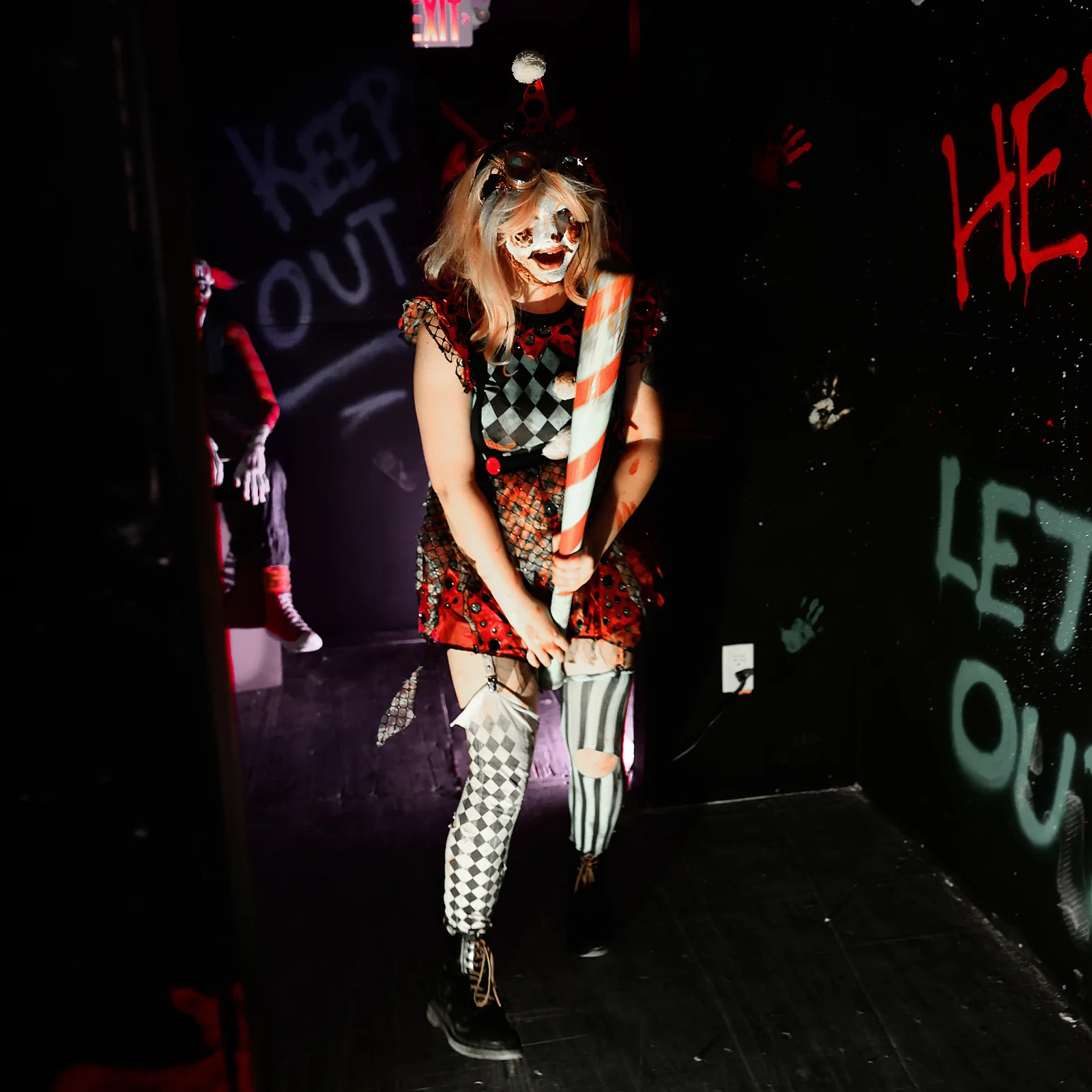 A clown performer waits in a dark, brightly painted haunt room.